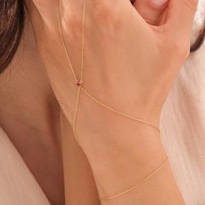 May include: A delicate gold hand chain featuring a thin, fine-link chain that wraps around the wrist and connects to a finger loop. A small, round, vibrant red gemstone is set at the center point where the chains meet on the back of the hand.
