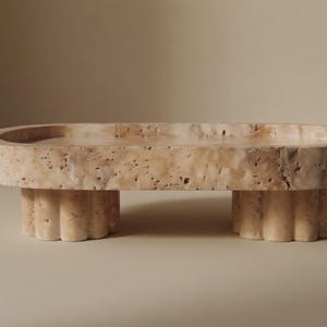 May include: A beige, oval travertine stone tray with a textured surface. It is supported by two fluted, cylindrical legs. The design is minimalist and modern, suitable for home decor.
