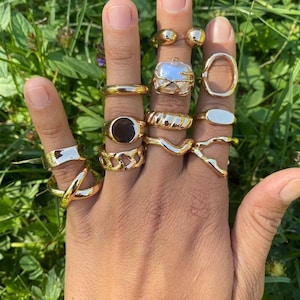 Peut inclure: Une collection de bagues dorées portées sur les doigts, présentant divers designs incluant une bague centrée sur une perle, un anneau à maillons, une chevalière ovale lisse, un anneau torsadé, une bague croisée et plusieurs anneaux polis.