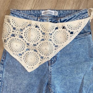 Peut inclure: Un jean en denim bleu clair délavé à taille haute avec un foulard triangulaire en crochet crème à motif circulaire drapé sur le devant. L'étiquette intérieure indique Stradivarius taille 38, USA 06, MEX 28, UK 10.