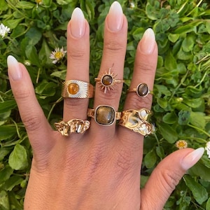 Peut inclure: Une collection de six bagues dorées portées sur les doigts. Les bagues présentent des designs variés, notamment un sertissage en forme de soleil avec une pierre brune, une pierre brune carrée, une pierre ovale lisse et des textures métalliques irrégulières.