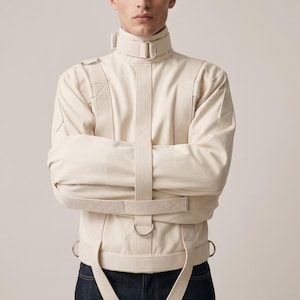 Könnte beinhalten: Cremefarbene Zwangsjacke mit mehreren Riemen und Metall-D-Ringen. Die Jacke hat einen hohen Kragen und wird über einem dunkelblauen Jeanshemd getragen. Das Kleidungsstück ist im Vintage-Stil.