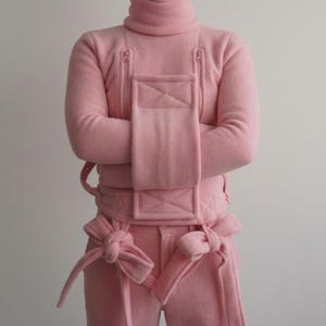 Op de afbeelding: Een lichtroze, eendelig outfit met een coltrui en bijpassende broek. Het kledingstuk heeft een gewatteerd borstpaneel, ritssluitingen en strikken bij de taille en enkels. De stof lijkt op een zacht, viltachtig materiaal. Een uniek mode-statement.