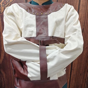 May include: A long-sleeved off-white garment featuring brown leather-like accents across the chest, shoulders, and hem. The piece includes a horizontal brown strap with a metal buckle across the midsection and a vertical strap extending downward.