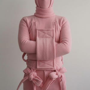 May include: A light pink, hooded jumpsuit with a high turtleneck, zippered chest pockets, and a waist belt. The jumpsuit features a unique design with bow details at the waist and on the pants. The model is wearing the jumpsuit with their arms crossed.
