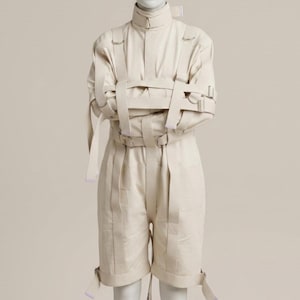 May include: A beige straightjacket featuring a high collar, long sleeves, and multiple adjustable fabric straps with metal buckles. The garment includes a central vertical strap, horizontal chest straps, and additional straps at the waist and cuffs.