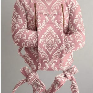 May include: A dusty pink baby carrier featuring a white damask pattern throughout. The carrier includes a high neck panel, two vertical brass-colored zippers on the front, and long fabric straps at the base for securing the carrier around the body.