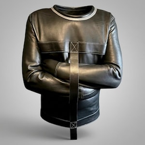 Op de afbeelding: Een zwart kledingstuk met lange mouwen en een ronde halslijn met een zilverkleurige bies. Het kledingstuk is voorzien van een horizontale borstband en een verticale middenband die naar beneden loopt, beide vastgezet met witte stiksels.