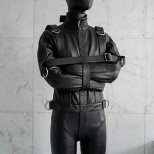 Può includere: Un indumento di contenzione in pelle nera con colletto alto, maniche lunghe e molteplici cinghie integrate con fibbie e anelli a D in metallo argentato. Il design presenta una cinghia centrale verticale e fasce orizzontali sul petto e in vita.