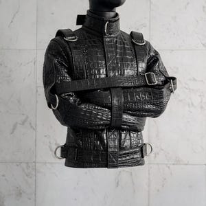 May include: A black jacket with a textured crocodile skin pattern, featuring a high collar with a metal buckle, multiple horizontal and vertical straps with silver-toned metal D-rings, and adjustable buckles across the chest and waist area.