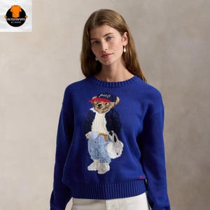 Puede incluir: Un suéter azul real con un diseño de Polo Bear. El oso lleva una chaqueta azul marino, una gorra roja y vaqueros azules. El suéter tiene cuello redondo y puños acanalados. La palabra "Polo" está bordada en el suéter.