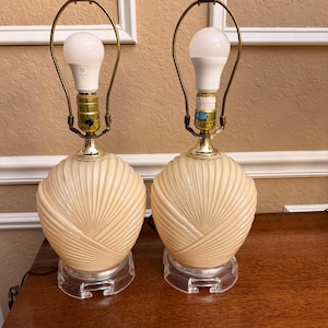 May include: A pair of table lamps featuring cream-colored, ribbed glass bases with a fan-like pattern. Each lamp has a gold-toned metal neck, a brass-colored socket with a white light bulb, a metal harp, and a clear, circular acrylic base.