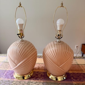 May include: A pair of table lamps featuring spherical, pale pink glass bodies with a raised, swirling ribbed pattern. Each lamp sits on a polished gold-tone metal base and includes a matching gold-tone metal harp fixture with a white light bulb.