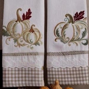 Może przedstawiać: Dwie białe ściereczki kuchenne z haftowanymi wzorami dyń w odcieniach złota, zieleni i burgundu. Każda ściereczka posiada ozdobną białą koronkę oraz dolny pas z beżowo-białej tkaniny w kratkę z akcentem w postaci wstążki w kropki.