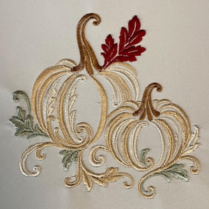 May include: An embroidery design featuring two pumpkins rendered in gold and tan thread with intricate swirling vine details. The larger pumpkin has a deep red leaf accent at the top, while smaller sage green leaves are positioned at the base.