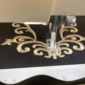 Puede incluir: Una aguja de máquina de coser y un prensatelas de plástico transparente se sitúan sobre una tela negra con un diseño bordado de volutas doradas. Se ve un bastidor de bordado blanco en la parte inferior con el texto 360x200 en relieve.