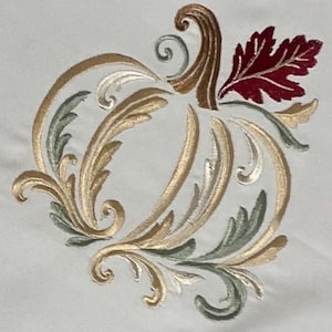 May include: An embroidered design on white fabric featuring a stylized pumpkin shape. The pumpkin is composed of swirling gold and sage green acanthus-style leaves, with a brown stem and a single deep red leaf accent at the top right corner.