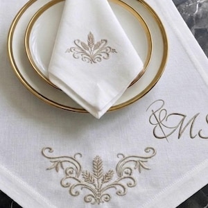 Pode incluir: Um conjunto de guardanapo e lugar americano em linho branco com bordado em fio bege de um motivo de volutas inspirado no trigo. O lugar americano inclui as letras R e M bordadas em cursivo. Um prato branco com borda dourada está posicionado.