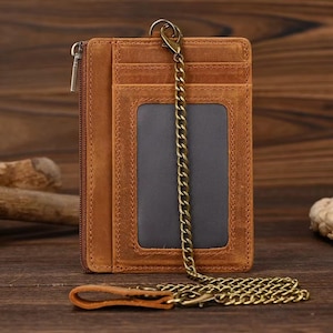Puede incluir: Una cartera de cuero marrón con cierre de cremallera, múltiples ranuras para tarjetas y una ventana de identificación transparente. Cuenta con una cadena de metal con un cierre y un pequeño lazo de cuero. La cartera es de cuero texturizado.