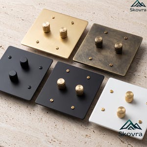May include: Five square light switches with two cylindrical knobs each. The collection includes a brushed gold plate, an aged brass plate, a matte black plate with black knobs, a matte black plate with gold knobs, and a white plate with gold knobs.