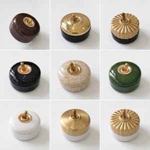 May include: A collection of nine round light switches featuring a central brass toggle lever. The switches vary in finish, including glossy brown, matte black with a gold cap, fluted gold, solid black, cream crackle, forest green, white, and gold.