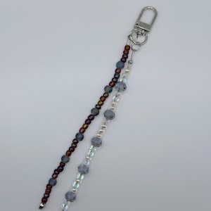 May include: A beaded keychain with two strands. One strand features dark red and brown beads, while the other has clear and gray beads. A silver-colored clasp and rings are at the top.