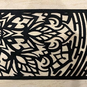 May include: A rectangular black laser-cut wooden panel with intricate designs. The panel features a floral mandala pattern on the left, a central ornamental design, and a vertical striped pattern on the right. The text "R-01" is in the upper left corner.