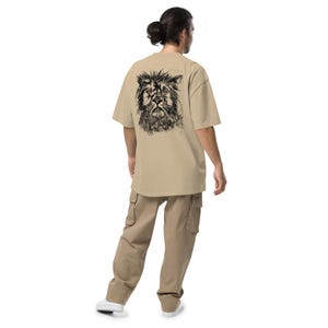 Puede incluir: Una camiseta color arena con un boceto de león negro en la espalda. Se combina con pantalones cargo color arena. El diseño del león está detallado con una apariencia texturizada y dibujada a mano. El modelo lleva zapatillas blancas.