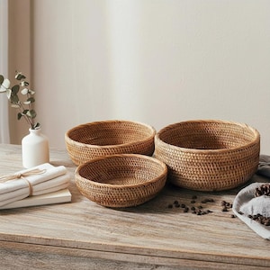 Puede incluir: Tres cuencos de ratán tejido de diferentes tamaños sobre una superficie de madera. Los cuencos son de color marrón natural cálido y tienen un aspecto texturizado. El cuenco más grande tiene unos 30 cm de diámetro, el mediano 25 cm y el más pequeño 20 cm.