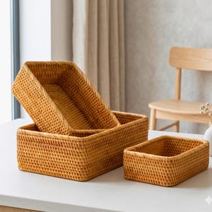May include: Three rectangular woven rattan baskets in varying sizes. The baskets are a warm, natural tan colour and have a textured surface. The largest basket is partially stacked on top of another, with a third basket to the right.