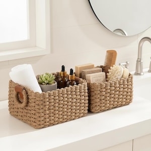 Può includere: Due cesti rettangolari intrecciati con vari articoli da bagno. Un cesto contiene un asciugamano bianco arrotolato, una piccola pianta grassa e bottiglie marroni. L'altro cesto contiene saponette, un pettine di legno e una spazzola per il corpo. Entrambi i cesti hanno manici in pelle marrone.