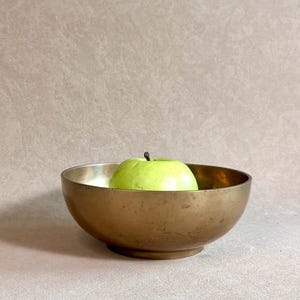 Può includere: Una ciotola rotonda in ottone metallico con una finitura liscia e leggermente usurata e un'apertura ampia. All'interno della ciotola si trova una mela intera di colore verde chiaro con un piccolo picciolo marrone, in contrasto con il metallo.