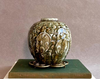 May include: A decorative, olive green ceramic vase with a detailed floral pattern, resting on a gold-toned metal stand. The vase sits atop two books, one green and one white, with the titles "LA TRAVIATA" and "LEONARDO DA VINCI" visible.