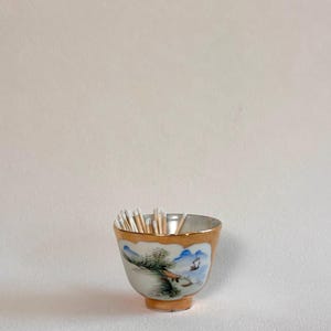 May include: A small, decorative ceramic bowl with a gold rim and a painted landscape scene. The bowl contains several wooden matches. The landscape features trees, mountains, and a small boat. The bowl is likely used for holding matches or small items.