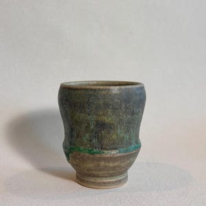 May include: A ceramic cup with a tapered base and a wide rim. The upper portion features a mottled dark green and charcoal glaze, while the lower section has a textured, earthy brown finish. A vibrant green glaze accent line separates the two colors.