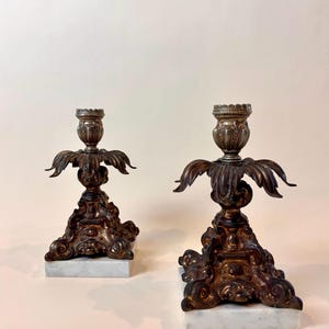 May include: Two antique bronze candle holders with ornate detailing. Each holder features a decorative base atop a white marble block. The candle holders have a detailed floral design and are suitable for home decor.