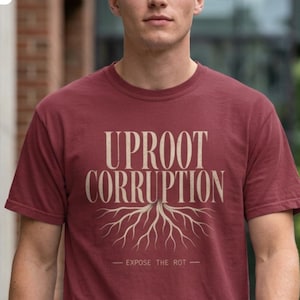 May include: A maroon short-sleeved t-shirt featuring the text UPROOT CORRUPTION in a light tan serif font above a graphic of tree roots. Below the roots, the text EXPOSE THE ROT is printed in a smaller font. The shirt has a crew neckline.