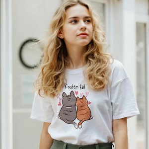 Peut inclure: T-shirt blanc avec un motif de deux chats enlacés et le texte "Foster fail". Les chats sont gris et orange, avec des coeurs roses. Le t-shirt est porté avec un pantalon vert.