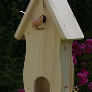 Könnte beinhalten: Ein helles Vogelhaus aus Holz mit einem dekorativen Vogel, der auf einem kleinen Ast sitzt. Das Vogelhaus hat ein Satteldach und einen abgerundeten Sockel. Der Vogel ist in Rosa- und Brauntönen bemalt.