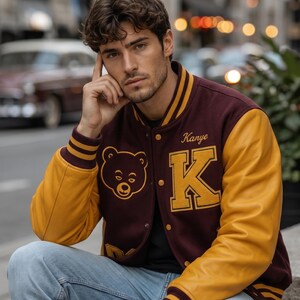 Puede incluir: Chaqueta universitaria granate y dorada con el nombre "Kanye" y un gran parche "K". La chaqueta tiene mangas doradas y un parche de oso. La persona lleva vaqueros azules.
