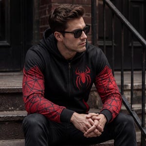 May include: Black zip-up hoodie with red spiderweb patterned sleeves and a red spider emblem on the chest. The hoodie has a hood and is worn with black sunglasses and black pants. The person is sitting on outdoor steps.