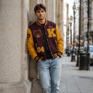 Puede incluir: Una chaqueta universitaria granate y dorada con el nombre "Kanye" y la letra "K" en la parte delantera. La chaqueta tambi&eacute;n presenta un gr&aacute;fico de oso y el n&uacute;mero "04". La persona lleva una camiseta negra, vaqueros azul claro y zapatillas blancas.