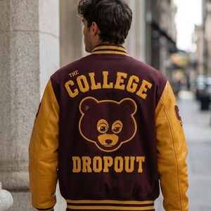 Puede incluir: Chaqueta universitaria granate y dorada con el texto "THE COLLEGE DROPOUT" y un gr&aacute;fico de oso en la espalda. La chaqueta tiene mangas doradas y pu&ntilde;os y dobladillo a rayas. La persona lleva vaqueros azul claro.