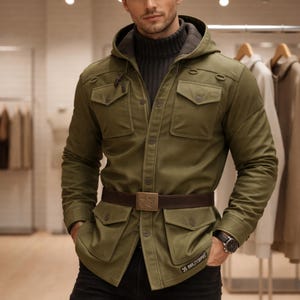 Può includere: Giacca verde oliva con cappuccio, quattro tasche anteriori e cintura marrone con fibbia a forma di stella. La giacca è indossata sopra un dolcevita grigio e jeans neri. Un orologio da polso è visibile sul polso sinistro.