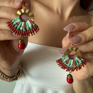 May include: A pair of handmade earrings with a gold-tone circular top and a fan-shaped design. The earrings feature red, turquoise, and brown woven threads, with a red teardrop-shaped bead hanging from the bottom. The earrings are held up by a person.