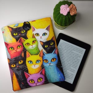 Puede incluir: Una colorida funda para lector electrónico con una vibrante ilustración de múltiples gatos con varios colores de ojos. La funda es amarilla con un cierre de botón negro. También hay un lector electrónico negro y un pequeño cactus verde con flores rosas y melocotón.