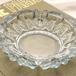 May include: Clear glass ashtray with a decorative, geometric pattern around the rim. The ashtray has a round shape with a central depression and is sitting on a book with gold lettering.