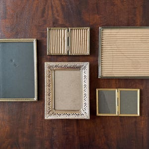 May include: A collection of vintage picture frames in various shapes and sizes. The frames are gold and silver, with some featuring ornate detailing. The backgrounds are neutral, with some having a corrugated texture. The frames are arranged on a dark wood surface.