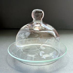 May include: A clear glass cloche with a round knob on top, resting on a matching plate. The glass dome features etched floral designs. The plate has a slightly raised edge. Ideal for serving cheese or desserts.