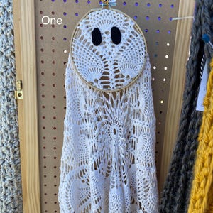 May include: A handmade white crochet ghost decoration with black oval eyes, framed in a wooden embroidery hoop. The ghost is hanging on a pegboard, with the word "One" visible. Other crochet items are in the background.